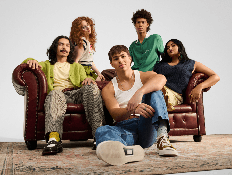 A group of five young people around a brown leather couch, some wearing shades of green and yellow