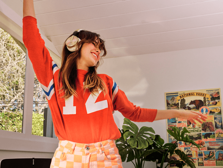 Woman in coral shirt and checkered salmon-colored pants enjoying music through headphones