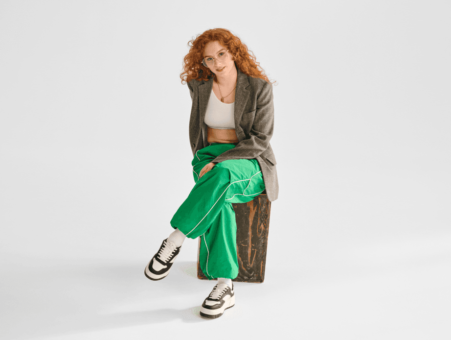Red haired woman wearing a white crop top, light brown blazer, and bright green track pants