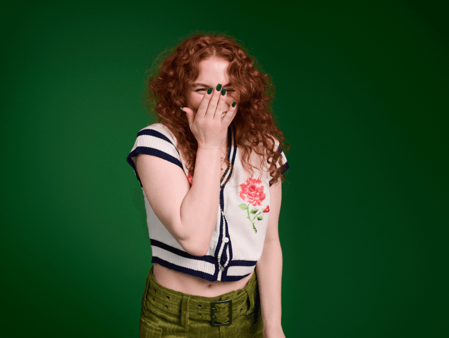 Red haired woman laughing as she covers her face, wearing dark green nail polish, a white short-sleeve sweater, and green pants against a green background 