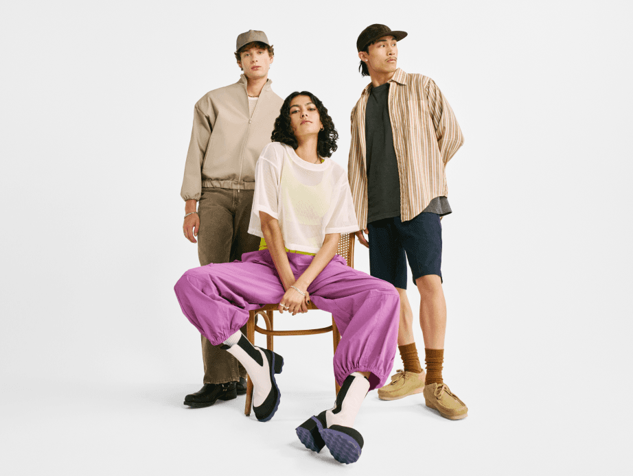 Two young people wearing neutral tones standing behind a person wearing a sheer white shirt over a neon yellow sports bra and bright lilac pants