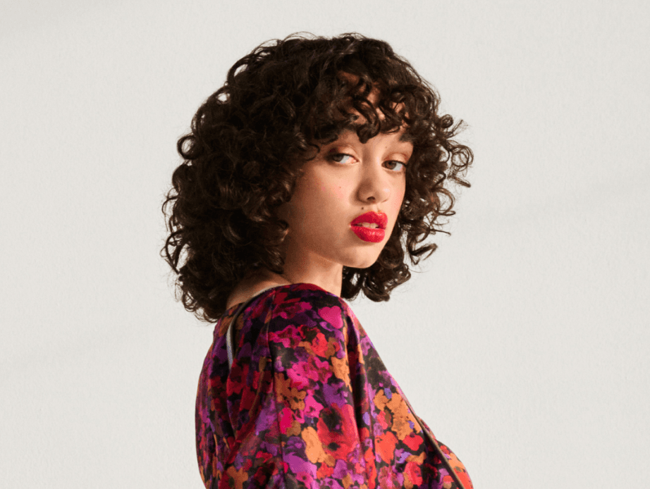 A woman with vibrant red lipstick and colorful pink, orange, and purple flower patterned dress against a light grey background