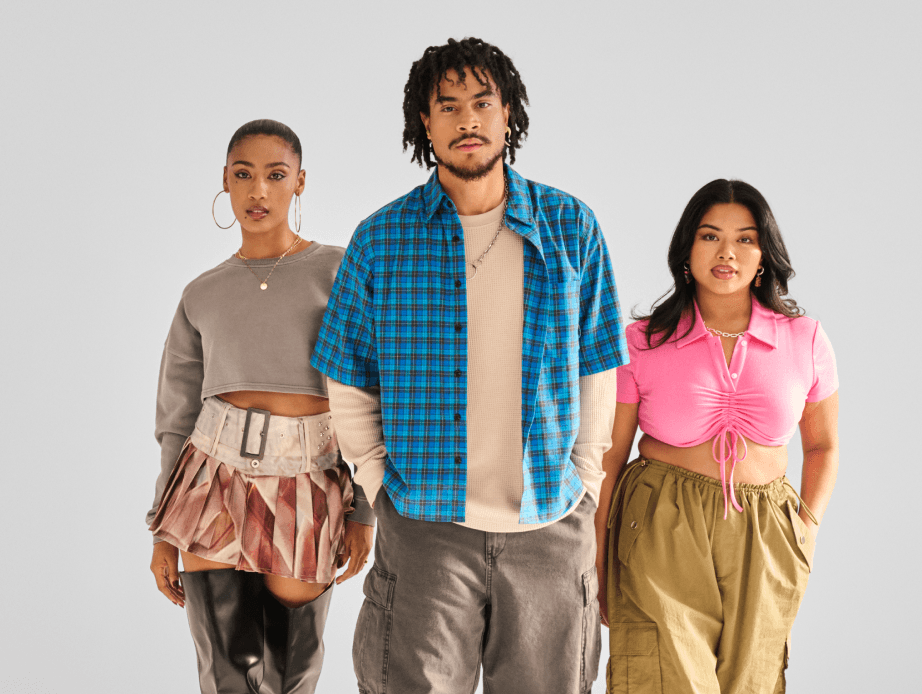 Three young people: one in a neutral long-sleeve sweater, one in a pink cropped polo shirt, and the third in an open blue plaid buttonup layered over a neutral base shirt