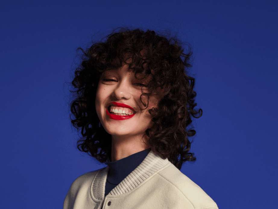 A woman with vibrant red lipstick, a dark violet mock neck, and a white varsity jacket smiling in front of a deep violet background